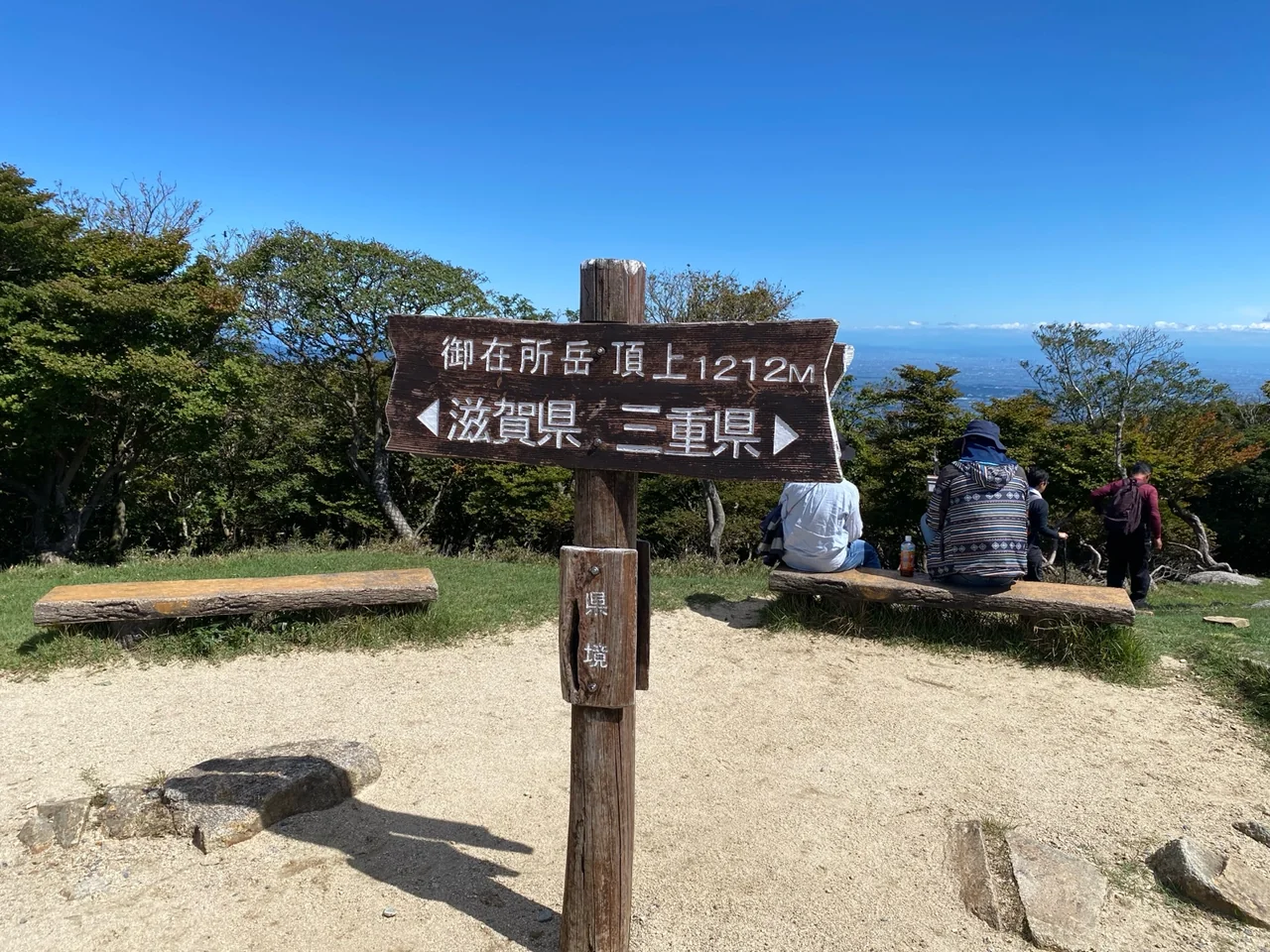 青空を背景にした御在所岳山頂の標識と、休憩する登山者